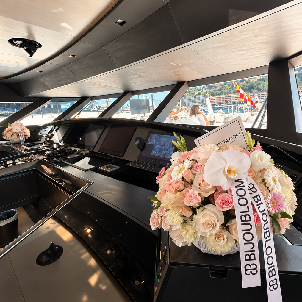 A luxury yacht bridge interior with a large floral arrangement of pink, peach, and white flowers on the console table. The bouquet features a white orchid and a white ribbon printed with the text  BIJOUBLOOM.